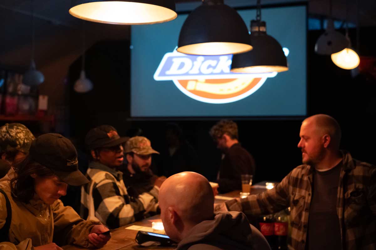 People crowded round a busy bar with a large projector screen in background with dickies logo on it.