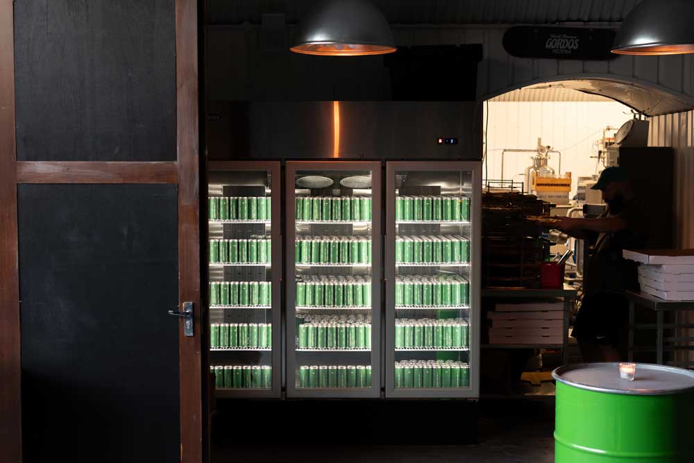 A large glass fridge full of green Umbrella London Apple Cider Cans with a man making fresh pizza in the background