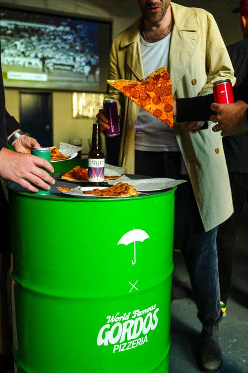 Men stood around a green barrel eating fresh pizza and drinking Umbrella London Ginger Beer